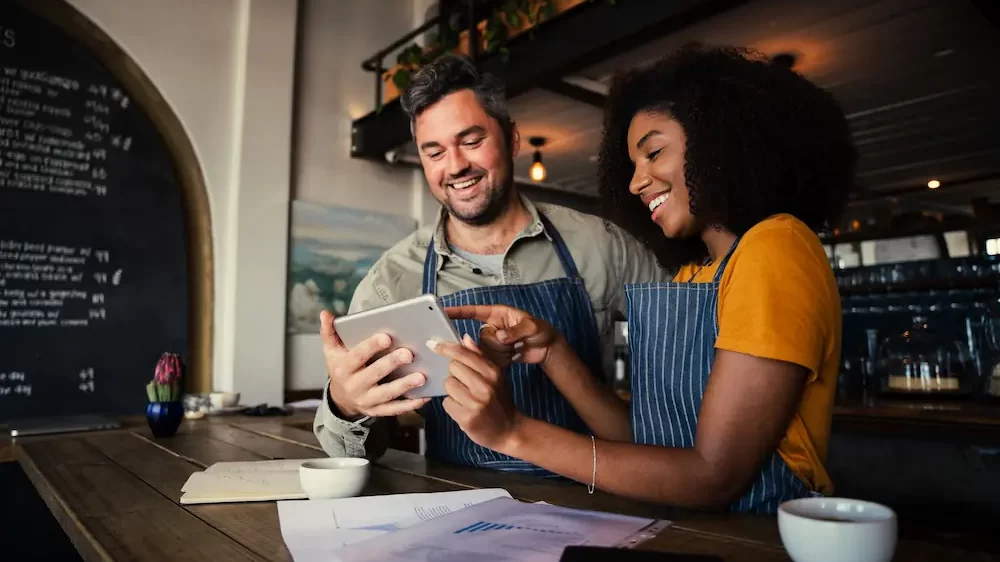 Manager and waitress laughing at designs on digital tablet standing with hot coffee in cafe