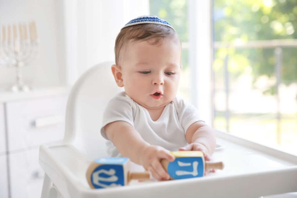 Cute baby in kippah playing with dreidels while sitting on high chair