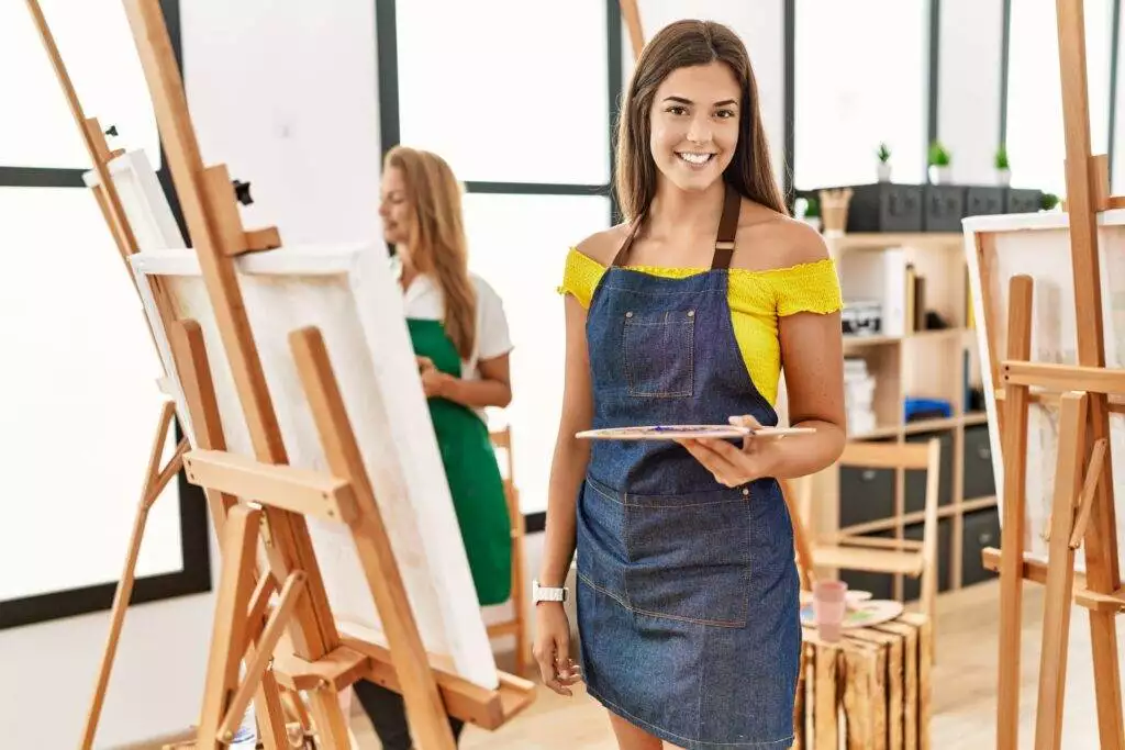 Young hispanic woman at art classroom looking positive and happy standing and smiling with a confident smile showing teeth