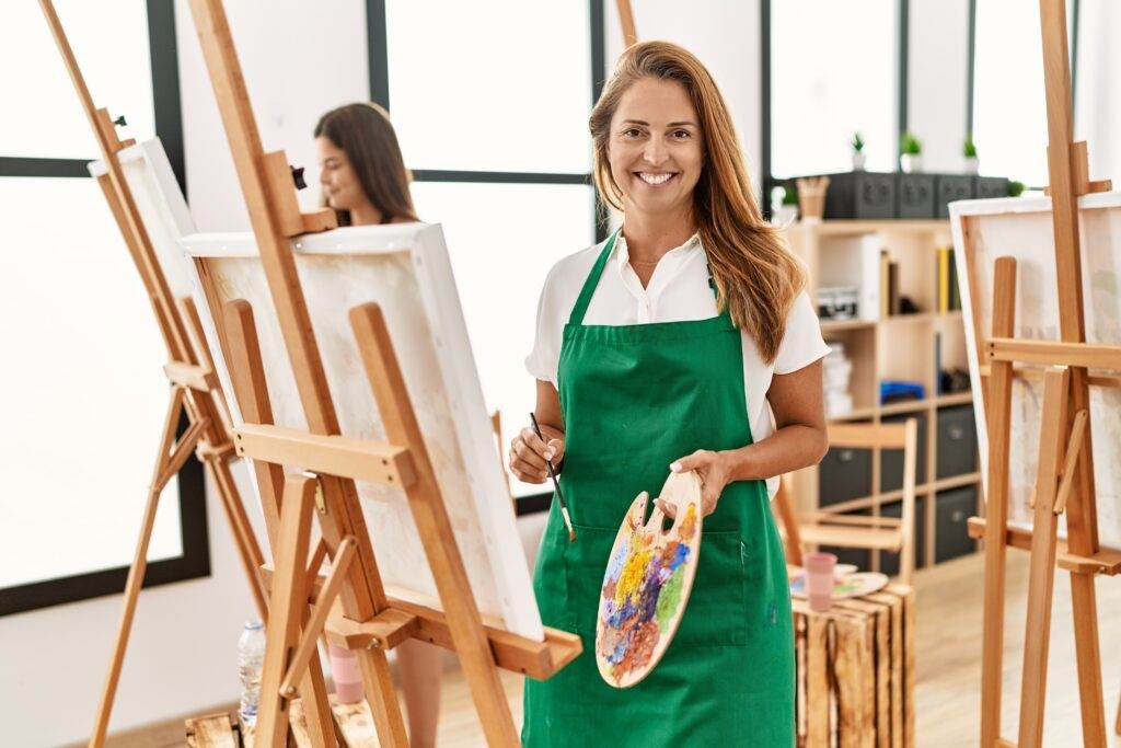 Young hispanic middle age woman at art classroom looking positive and happy standing and smiling with a confident smile showing teeth