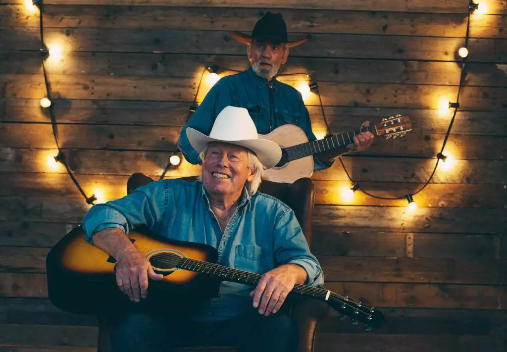 Two smiling senior country and western musicians sitting on chair in front of wooden wall with light bulbs.