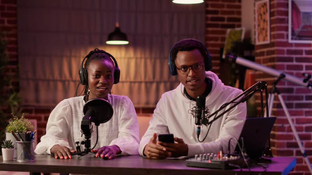 African american influencer couple streaming live dual vlog talking sitting at desk with podcast setup in home studio. Man and woman content creators recording smartphone review for social media.