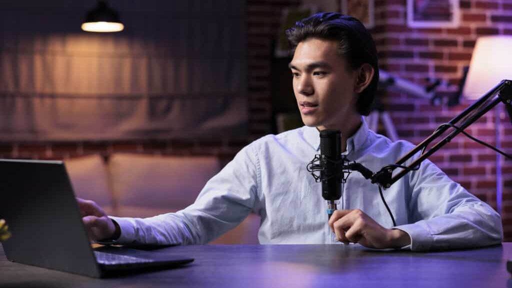 Asian blogger vlogging podcast show, live broadasting content on internet channel, using laptop and sound equipment. Recording video blog at audio station, talking to social media audience.