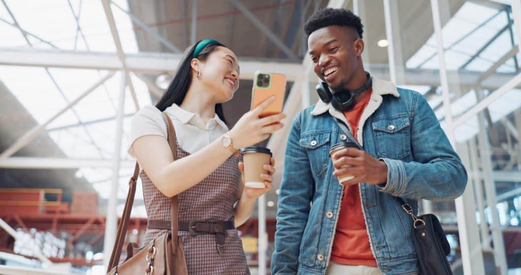 Shopping mall, smartphone and couple of friends on social media, website or blog for discount, sales and travel communication. Diversity gen z people walking, using phone or cellphone and coffee cup.