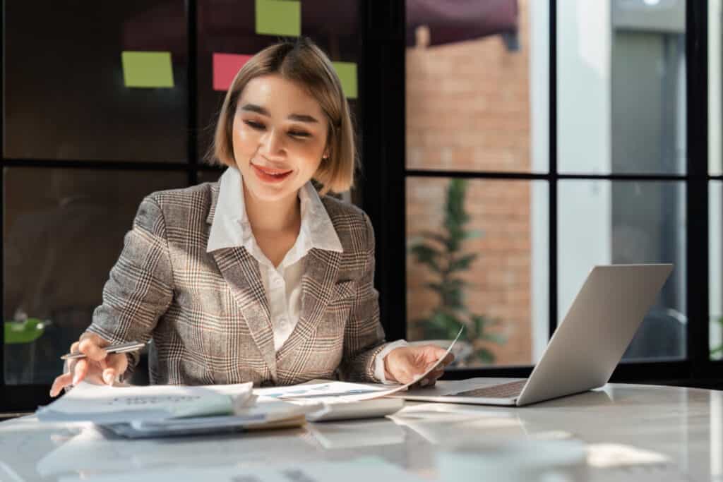 Young business woman holding accounting bookkeeping documents checking financial data or marketing report working in office with laptop. Paperwork manageme.