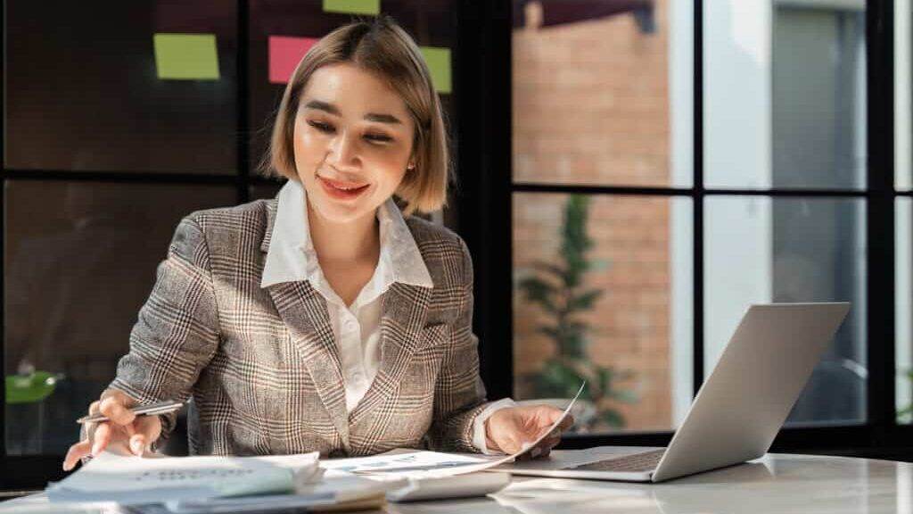 Young business woman holding accounting bookkeeping documents checking financial data or marketing report working in office with laptop. Paperwork manageme.