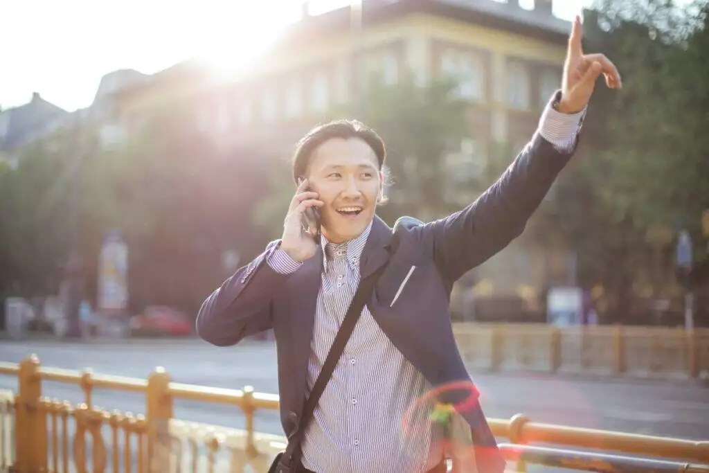 Happy adult ethnic man in jacket waving with hand hailing taxi on sunny street while speaking on smartphone