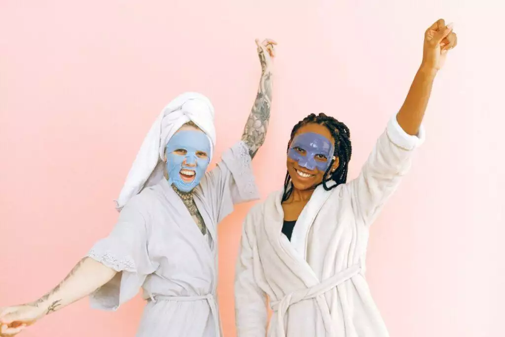 Happy diverse women in robes and blue collagen masks applied on face standing with raising hands near pale pink wall and looking at camera