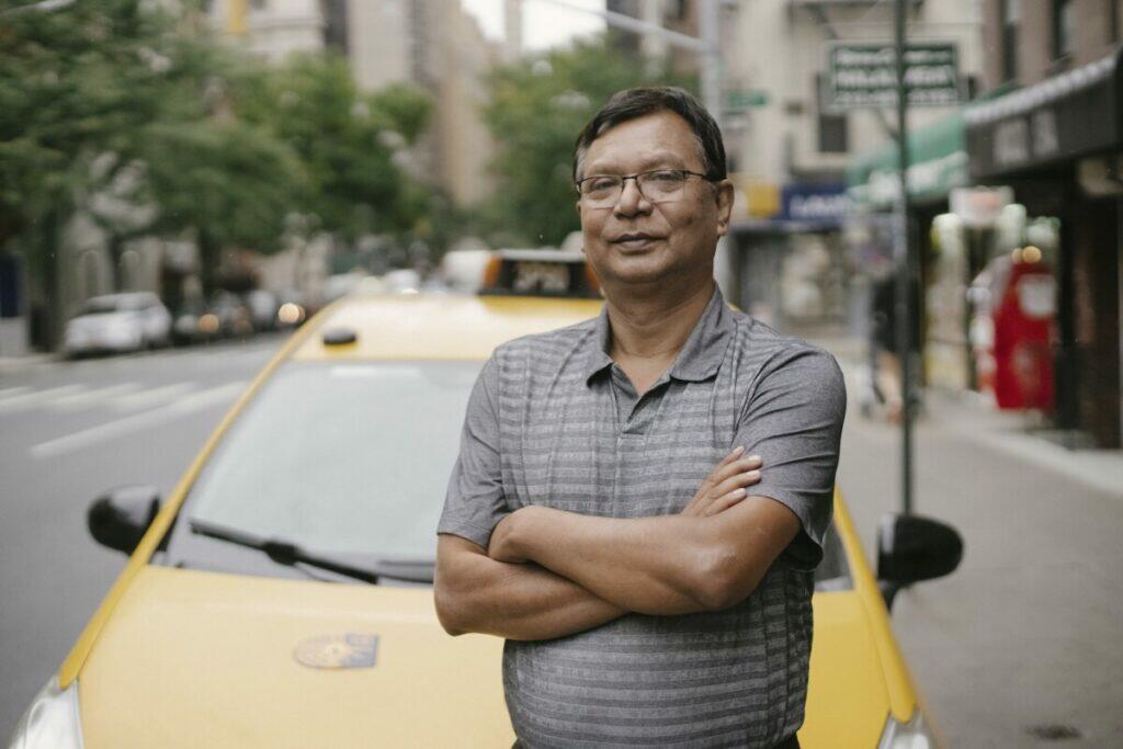 Confident Asian male in eyeglasses looking at camera while standing with crossed arms near yellow taxi on street with blurred background