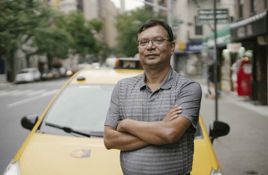 Confident Asian male in eyeglasses looking at camera while standing with crossed arms near yellow taxi on street with blurred background