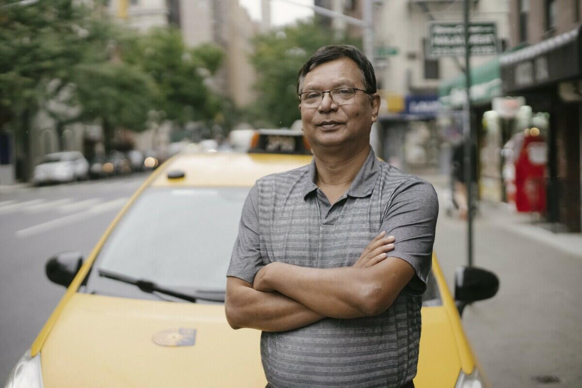 Confident Asian male in eyeglasses looking at camera while standing with crossed arms near yellow taxi on street with blurred background