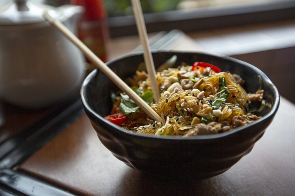 Delicious appetizing Asian noodles with vegetables and nuts served in bowl with chopsticks on table